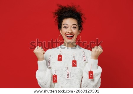 Young overjoyed excited fun happy female costumer woman 20s wear white knitted sweater with tags sale in store showroom do winner gesture clench fist isolated on plain red background studio portrait