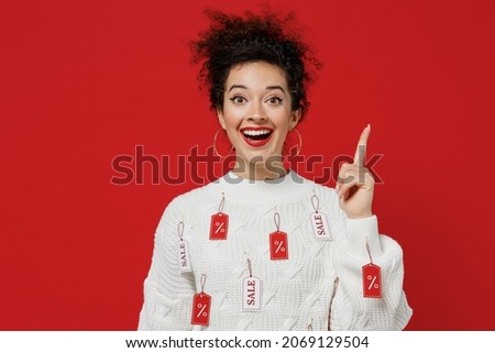 Young smiling happy female costumer woman 20s wear white knitted sweater with tags sale in store showroom holding index finger up with great new idea isolated on plain red background studio portrait
