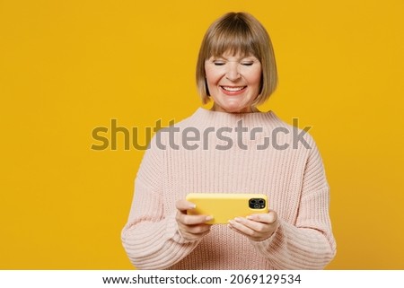 Elderly gambling woman 50s wears pink casual knitted sweater using play racing app on mobile cell phone hold gadget smartphone for pc video games isolated on plain yellow background studio portrait.
