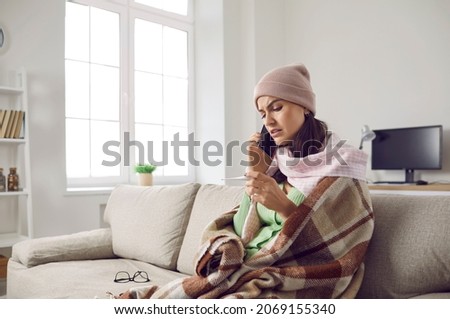 Unhappy woman who has flu or bad cold calling doctor. Sick young lady wearing warm hat, scarf and plaid sitting on sofa at home, looking at thermometer and making phone call to physician. Copy space Royalty-Free Stock Photo #2069155340