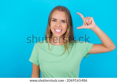 Beautiful caucasian girl wearing green T-shirt over isolated background smiling and confident gesturing with hand doing small size sign with fingers looking and the camera. Measure concept