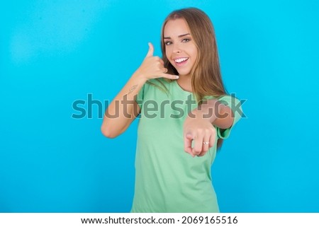 Beautiful caucasian girl wearing green T-shirt over isolated background smiling cheerfully and pointing to camera while making a call you later gesture, talking on phone