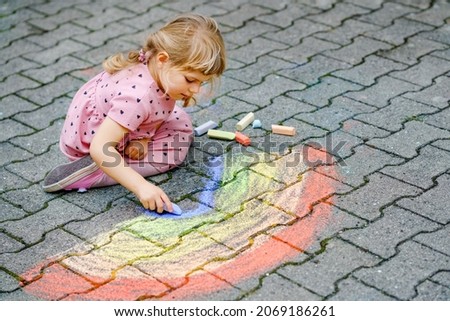 Little preschool girl painting rainbow with colorful chalks on ground on backyard. Positive happy toddler child drawing and creating pictures. Creative outdoors activity in summer.