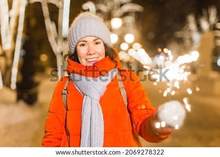 Smiling young woman wearing winter knitted clothes holding sparkler outdoors over snow background. Christmas holidays.