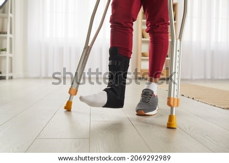 Unrecognizable person with broken leg or foot injury walking on crutches. Man wearing leg brace ankle support adjustable strap fracture fixator standing in living room. Cropped low section close up Royalty-Free Stock Photo #2069292989