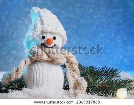 Cute knitted toy snowman in a hat and scarf in the snow, blue background. Christmas holiday decoration copy space