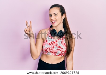 Young brunette teenager wearing gym clothes and using headphones showing and pointing up with fingers number two while smiling confident and happy. 