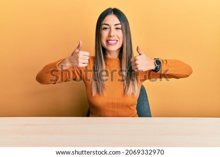 Young hispanic woman wearing casual clothes sitting on the table success sign doing positive gesture with hand, thumbs up smiling and happy. cheerful expression and winner gesture. 