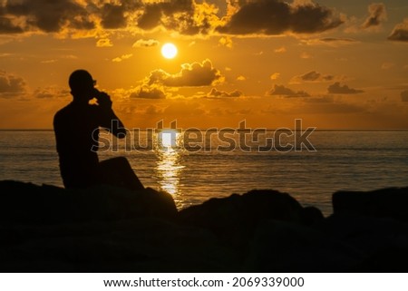 Silhouette of a man with glasses drinking coffee from a cup sitting on the seashore at sunset. Beautiful, magical sunset on the sea with a silhouette of a man. Photo from the back, focus on the sun