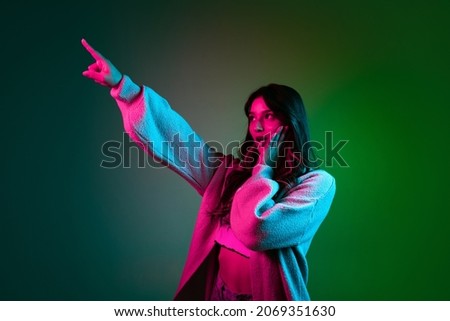 Pointed. Studio shot of young adorable cute girl, student in casual style clothes posing isolated on dark green studio backgroud in pink neon light. Emotions, facial expression, youth, fashion and ad