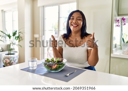 Young hispanic woman eating healthy salad at home success sign doing positive gesture with hand, thumbs up smiling and happy. cheerful expression and winner gesture.  Royalty-Free Stock Photo #2069351966