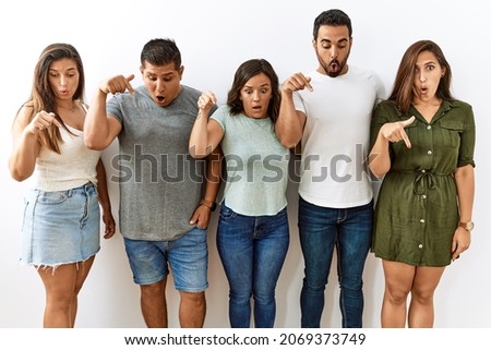 Group of young hispanic friends standing together over isolated background pointing down with fingers showing advertisement, surprised face and open mouth 