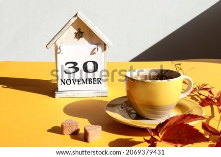 Calendar for November 30: a cup of tea with branches of girlish grapes against the background of a decorative house with the name of the month in English and the number 30, gray background
