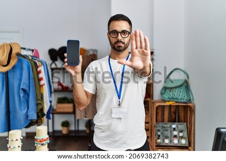 Young hispanic man with beard working as manager at retail boutique holding smartphone with open hand doing stop sign with serious and confident expression, defense gesture 