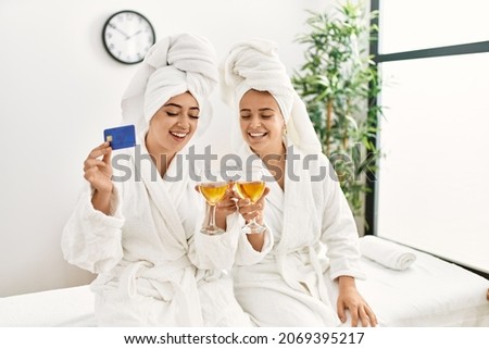 Woman couple holding credit card and toasting with champagne sitting on massage board at beauty center
