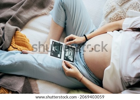 Unrecognizable pregnant woman in third trimester lying on bed looking at medical ultrasound scan. The concept of a healthy happy pregnancy and expectation of the birth of a baby. Royalty-Free Stock Photo #2069403059