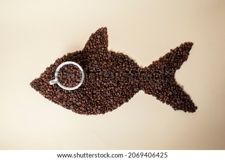 Cool fish made of caffee beans. Light Brown background. Eye cup of coffee.