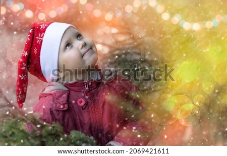 Christmas and new year banner with happy child girl in a red Santa hat waiting for a miracle. Laughing cute baby is on magical winter background with festive garlands lights, christmas tree and bokeh