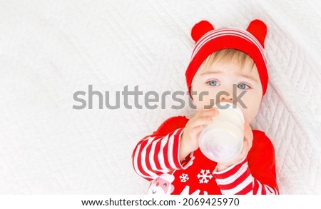 Little Caucasian baby boy wearing Christmas suit eating milk from bottle. First Christmas. Top view. White gray background. Banner, free space, copy space