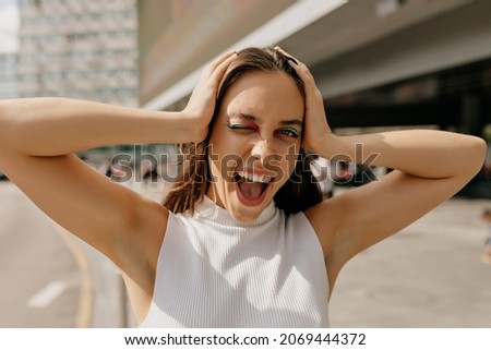 Carefree stylish excited woman with bright fashion make up holding hands on the head and screaming. Outdoor portrait of cute brunette girl on sunshine street.