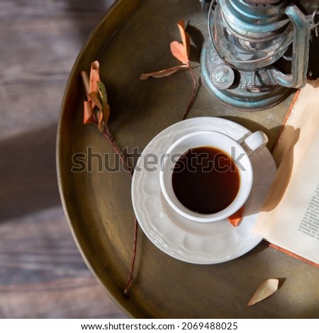 White cup of black coffee is on a metal table next to a book and a table lamp. Retro style. 
Square image.