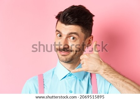 Close-up of handsome young guy with moustache, showing phone gesture, asking to call him, standing on pink background