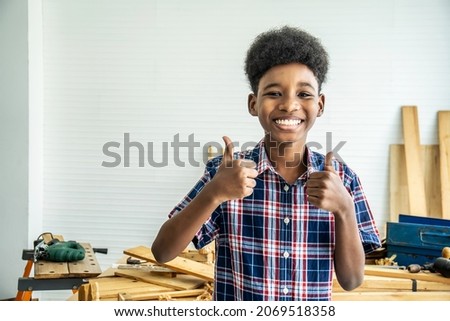 Smiling African-American boy carpenter standing with giving thumbs up as sign of success in a carpentry workshop.