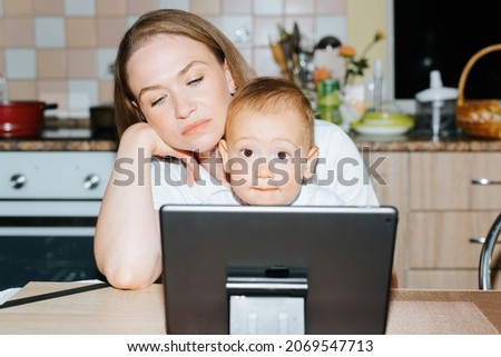 Tired mom and child using tablet watching educational video while sitting in kitchen at home. Little boy emotionally looking at camera. Motherhood, parenting, childcare concept.