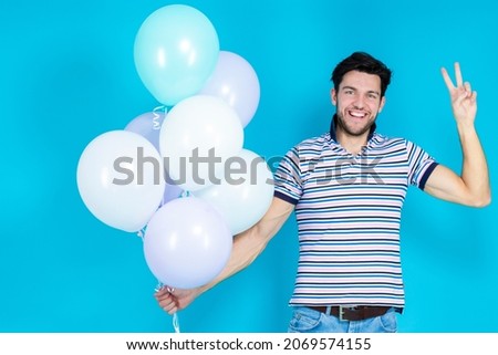 Celebration Ideas. Positive Tranquil Caucasian Handsome Brunet Man With Bunch of Colorful Air Balloons Posing With lifted Hand and V-Sign Against Blue Background. Horizontal Image