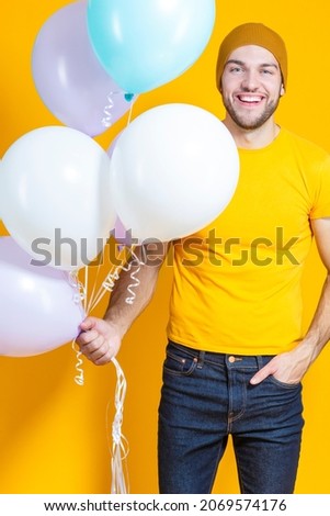 Funny Smiling Caucasian Young Handsome Man With Colorful Air Balloons Posing in Warm Hat Over Yellow Background.Vertical image