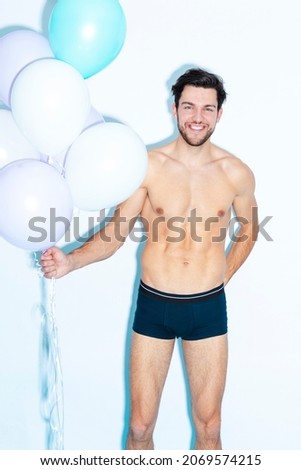 Funny Laughing Caucasian Handsome Brunet Man With Bunch of Colorful Air Balloons in Hand While Posing in Underware Against White Background. Vertical Image