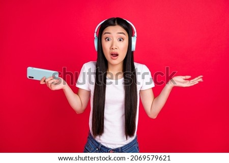 Photo of young asian girl unhappy shrug shoulders confused listen headphones hold cellphone isolated over red color background
