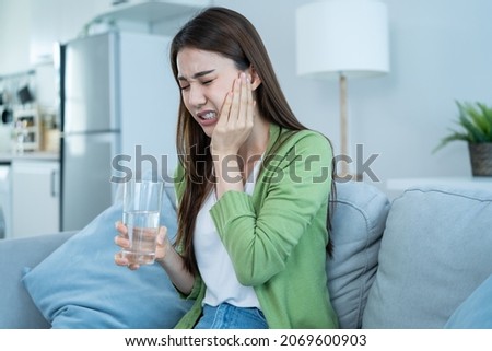 Asian beautiful woman feel terrible toothache after drink cold water. Attractive female sit on sofa in living room touching cheek, feel hurt and suffering from sensitive tooth ache, pain and cavities. Royalty-Free Stock Photo #2069600903