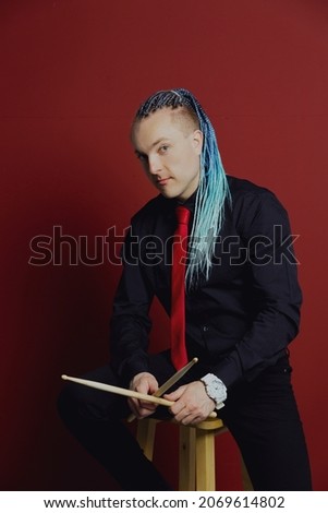 A young attractive man with an interesting hairstyle in pigtails on an undercut with a red tie on a red background in and drumsticks in his hands. Royalty-Free Stock Photo #2069614802