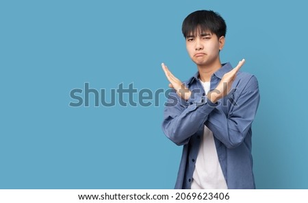 Portrait of young handsome Asian man showing STOP or Rejection expression crossing arms doing negative sign, angry face, isolated blue color background. Royalty-Free Stock Photo #2069623406