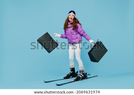 Full size skier woman in warm purple padded windbreaker jacket ski goggles mask spend weekend in mountains holding package bags with purchases after shopping isolated on plain blue background studio.