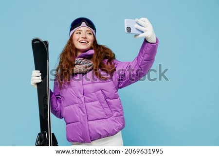 Skier woman in warm purple padded windbreaker jacket goggles mask spend extreme weekend in mountains doing selfie shot on mobile cell phone post photo hold ski isolated on plain blue background studio
