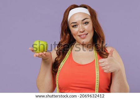 Young chubby overweight plus size big fat fit woman in red top measure tape warm up training eating green apple fruit show thumb up gesture isolated on purple background home gym Workout sport concept