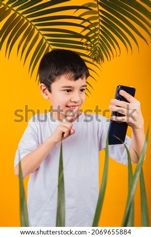 Portrait of Asian boy holding phone taking selfies showing v-sign on yellow background with palm leaves.