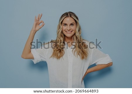 Relaxed young female showing ok gesture with hand and resting other one on her hip, assuring its alright and being confident it will all be fine while standing isolated next to blue wall