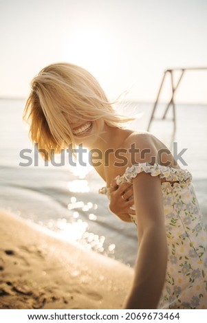 Pretty caucasian young woman on seashore bent over photo, smiling broadly. Her short blonde hair covers face, wearing sundress. Happiness summer lifestyle concept.
