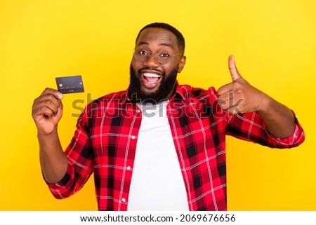 Photo of brown haired bearded amazed man wear red plaid shirt glasses thumb up credit card isolated on yellow color background