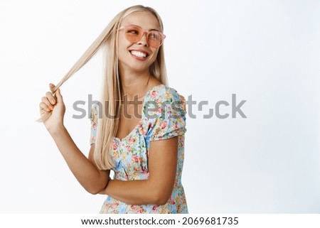 Beautiful blond woman in sunglasses, playing with hair, smiling and looking behind her at promo text, standing over white background