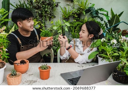 Young gardener woman wearing apron use smartphone take a photo her boyfriend, He holding house plant and smile with happy together
