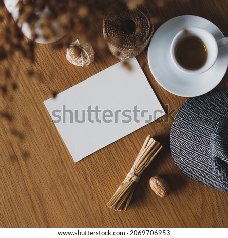 Autumn still life on wooden table in cafe. Blank paper card, coffee cup, female hat, dry grass in vase. Elegant feminine workspace, cozy home office desk table.