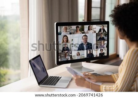 Concentrated young African American ethnic woman holding paper reports looking at computer monitor, holding video conference talk with diverse colleagues, discussing working issues in modern office.