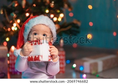 Portrait of cute baby in a Christmas hat holding a holidays gift. Festive lights and xmas gifts on the background. Merry Chrismas and Happy New Year concept.