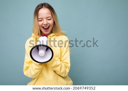 Portrait of beautiful pretty positive happy smiling blonde young woman with sincere emotions wearing hipster yellow hoodie isolated over blue background with copy space and shouting into loudspeaker
