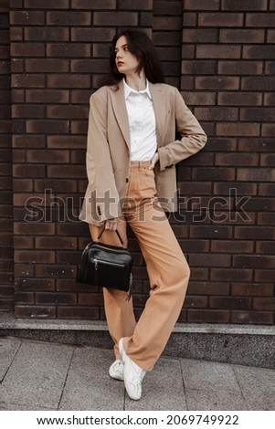 Tall stylish girl with a bag in a beige oversized jacket and orange pants on brick wall background Royalty-Free Stock Photo #2069749922