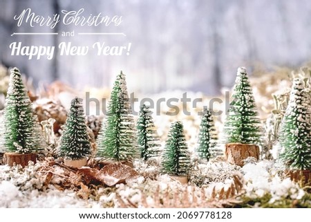 Christmas or New Year greeting card with group of decorative Christmas trees on snow covered moss with winter forest at background. Xmas holidays atmosphere. Greeting text include